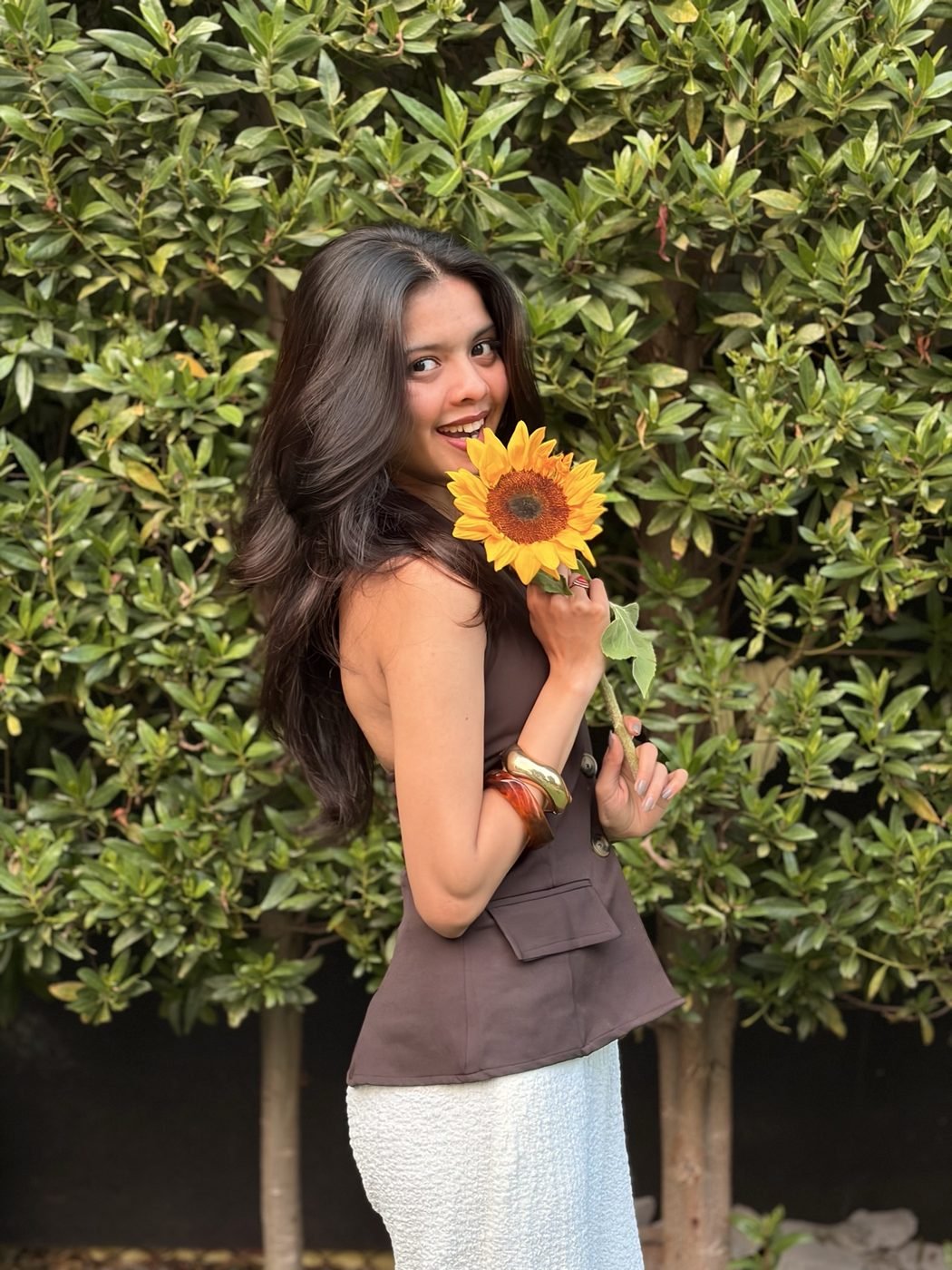 Disha Rokde holding a sunflower in front of a green hedge